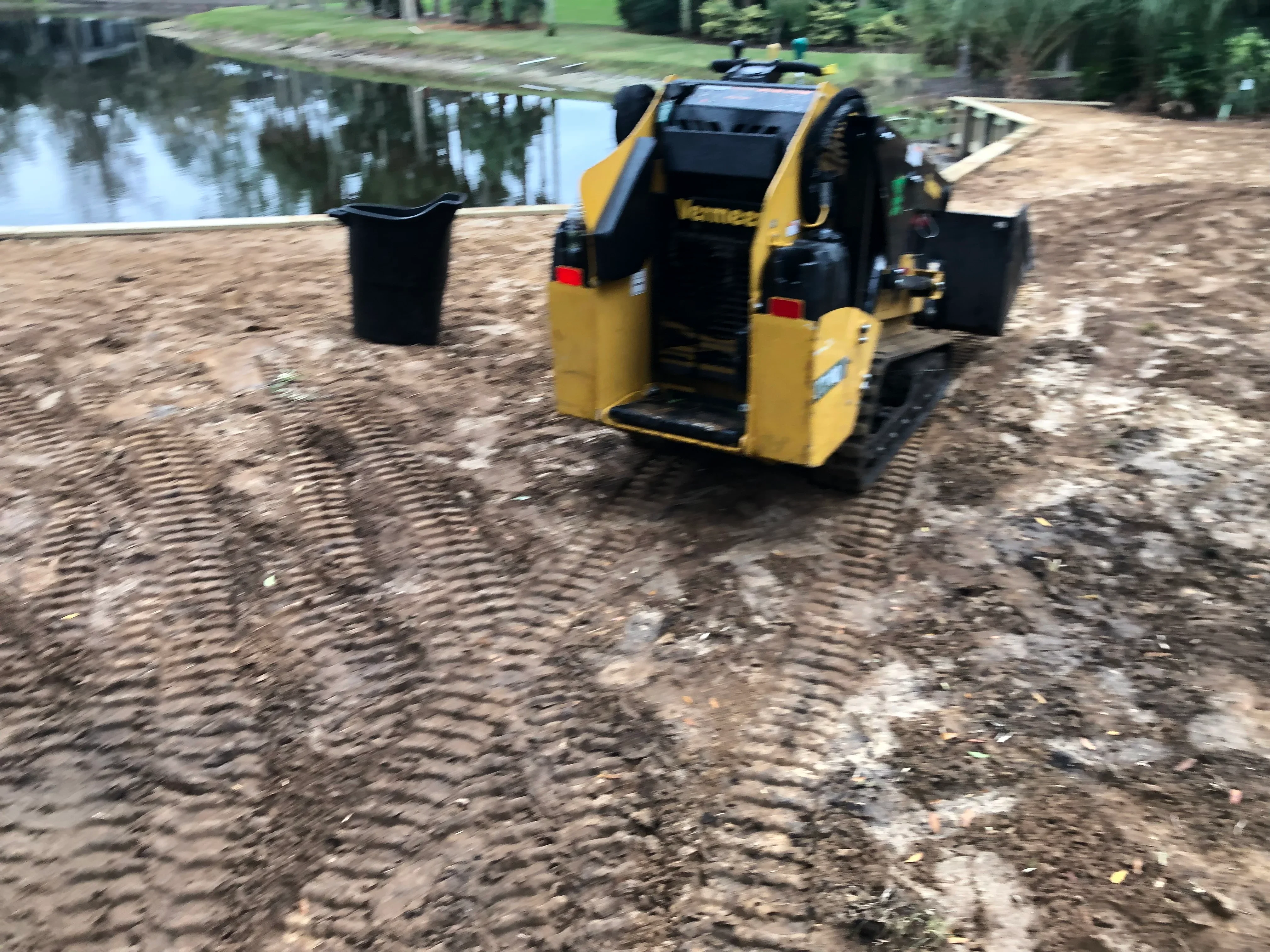 Yellow compact track loader grading dirt lot near water with black waste bins